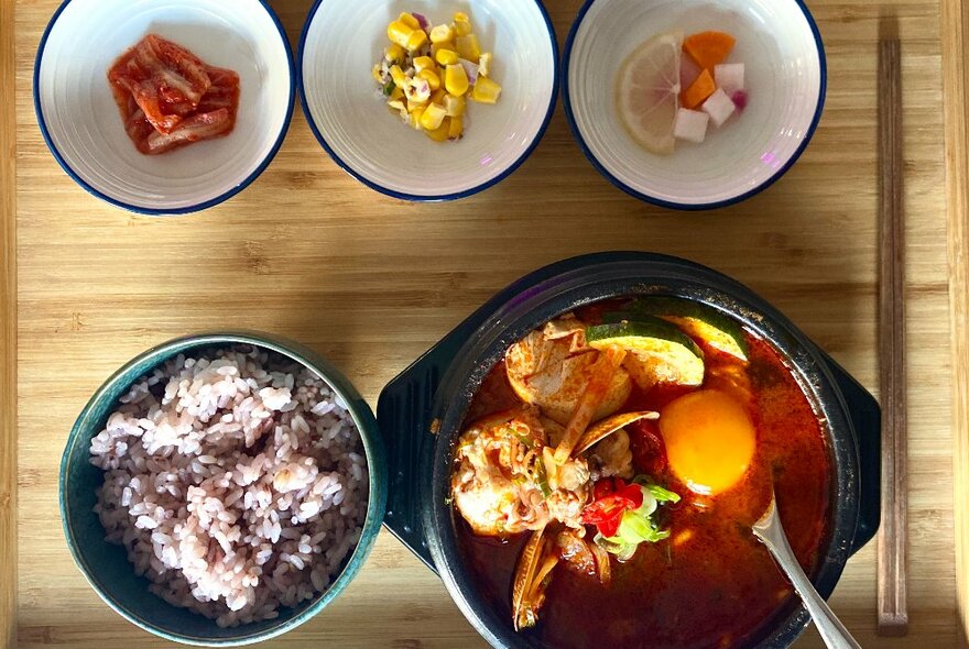 An authentic Korean hot pot in a green ceramic dish, with rice on the side and three small white bowls with garnishes to add, on a wooden serving board with fork and chopsticks.