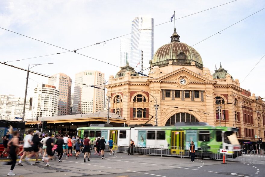 People running along the streets of Melbourne, past Flinders Street station and a tram, as part of an organised event.