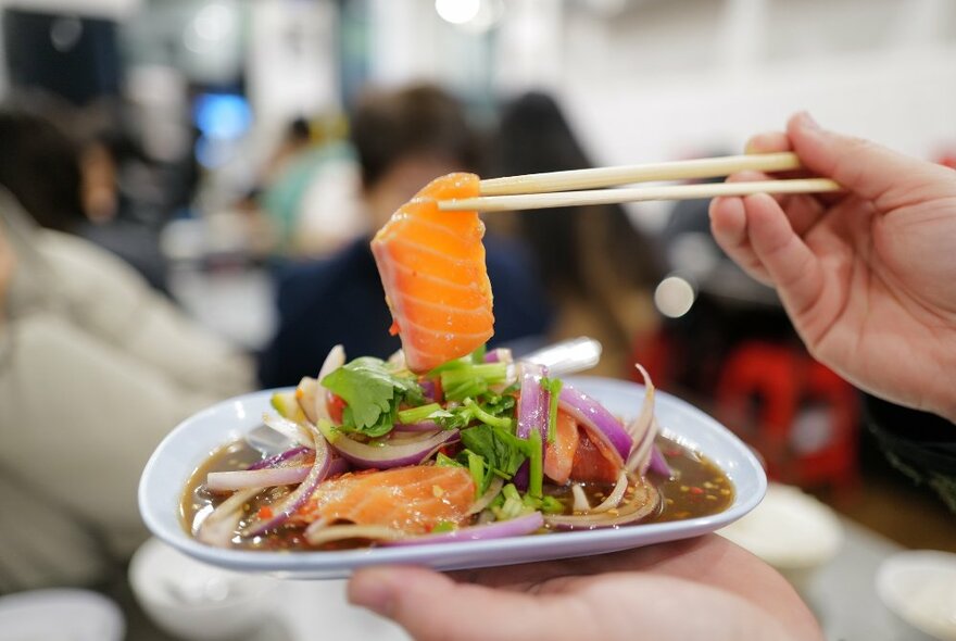 A hand holding chopsticks holding a slice of salmon above an Asian dish of food with gravy, greens and onions.