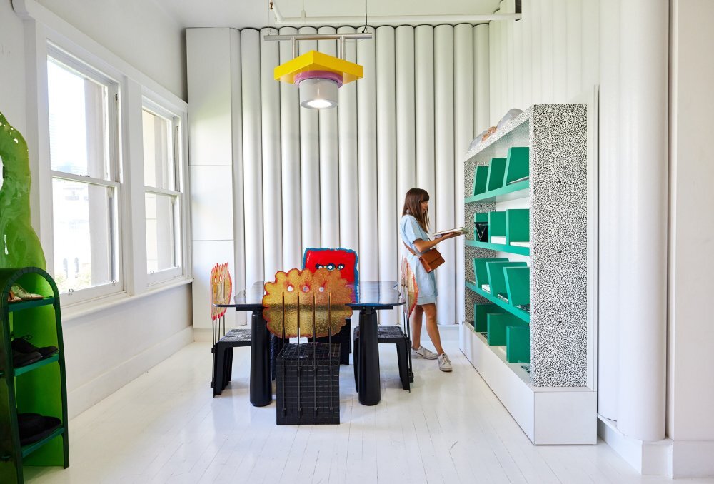 A woman is looking at books in a shop with white walls and flooring, and unique designer furniture.