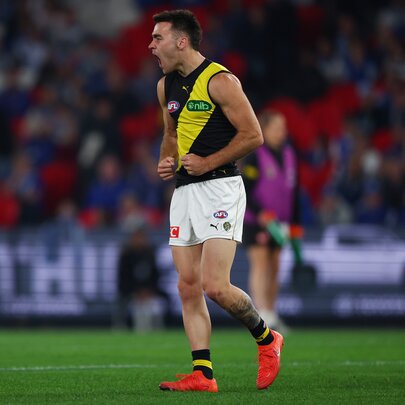 A Richmond AFL football player on the field during a match, a blurred crowd of stadium fans in the background.