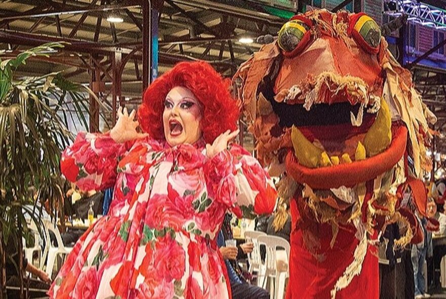 Drag performer wearing a curly red wig and red flowered dress screaming with hands raised next to a Chinese dragon in a market stall area.