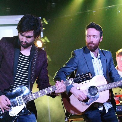 A band playing live on stage, two guitarists are in the foreground, with a drummer just visible behind them.