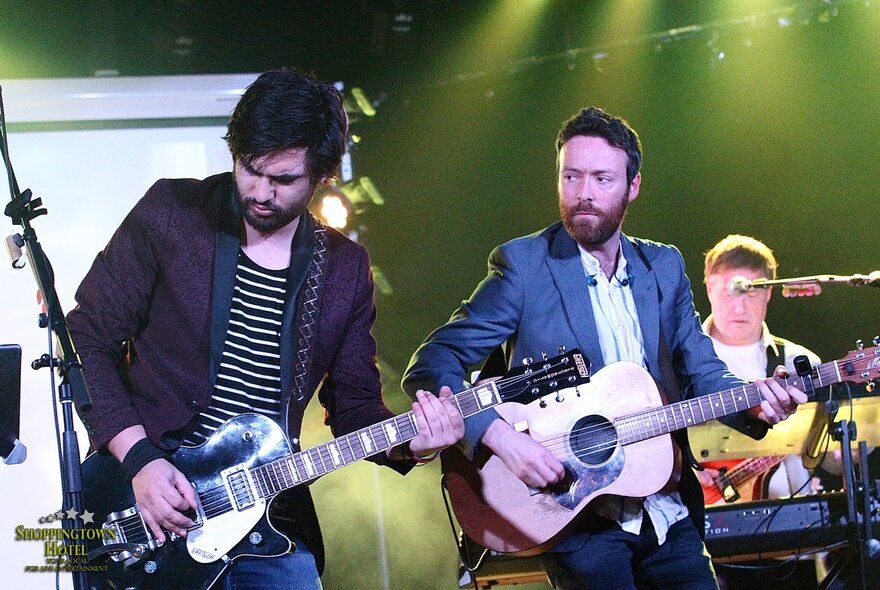 A band playing live on stage, two guitarists are in the foreground, with a drummer just visible behind them.