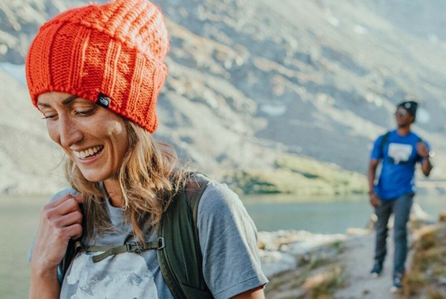 Smiling woman wearing orange thermal beanie on her head, walking outdoors near water with another walker in the distance behind her. 