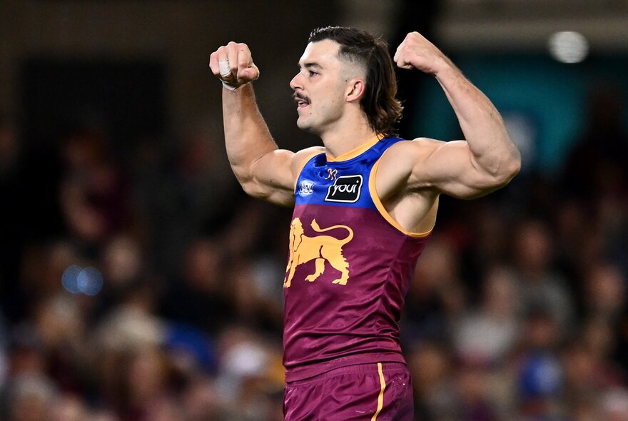 A Brisbane Lions AFL player celebrating on the field with both his fists clenched and his arms raised in a strong man position above his head.