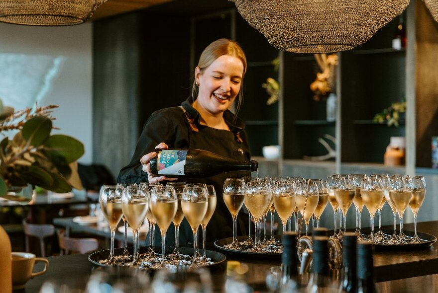 A waiter in a restaurant filling up trays of multiple flute-shaped glasses with sparkling wine.