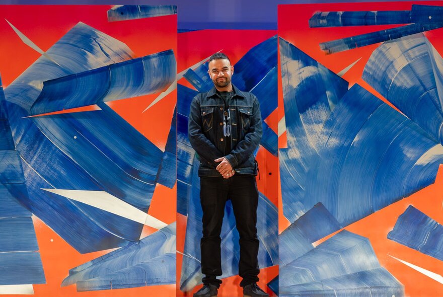 Artis Sahil Roy standing formally between two of his large paintings with large swathes of blue on and orange background