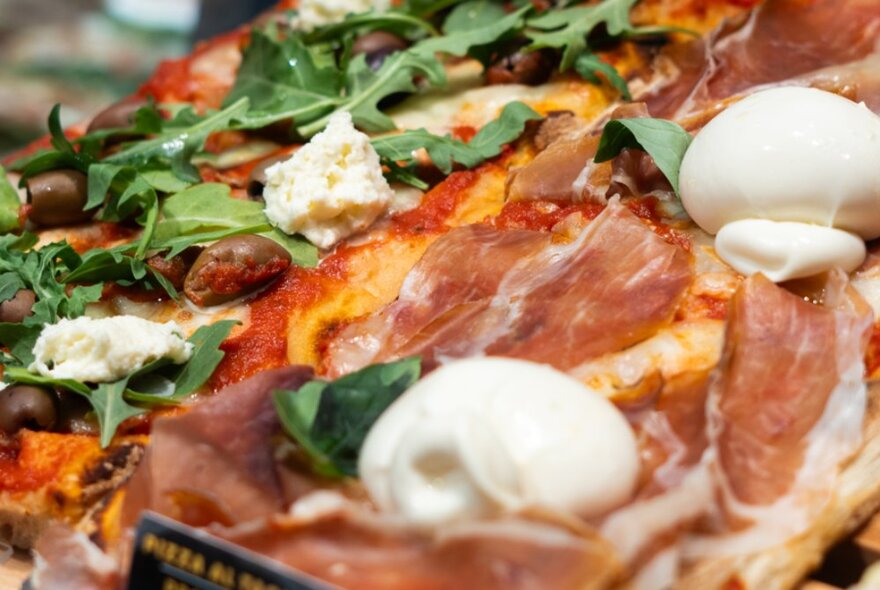 Close-up of a pizza with olives, thin slices of prosciutto, mozzarella and rocket topping.