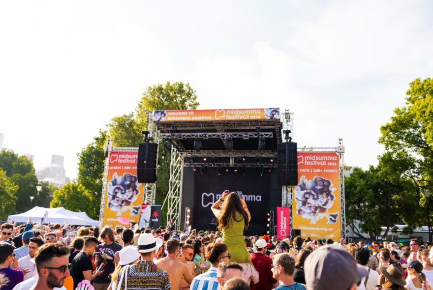 A crowd outdoors looking towards a stage, one person sitting on another's shoulders. 