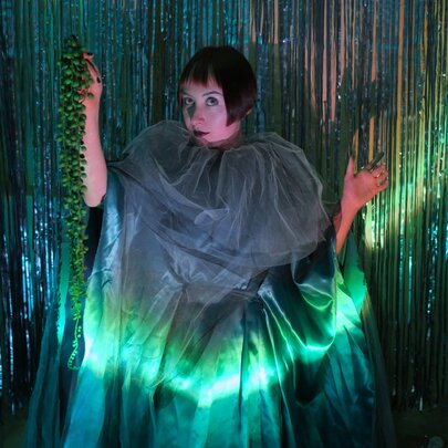 A performer wearing a dramatic sheer tulle green cape with a high gathered collar, illuminated by a vibrant green under-light against a tinsel backdrop.
