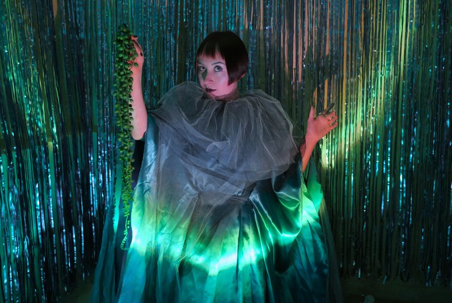 A performer wearing a dramatic sheer tulle green cape with a high gathered collar, illuminated by a vibrant green under-light against a tinsel backdrop.