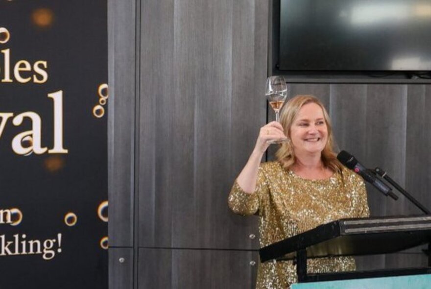 A smiling woman standing behind a lecturn holding up a glass of sparkling wine, behind her is a large sign with some letters just visible.