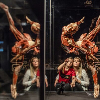 A woman and child sit in a museum, looking up at a glass case displaying a preserved human body posed in a dramatic leap, with exposed muscles visible.