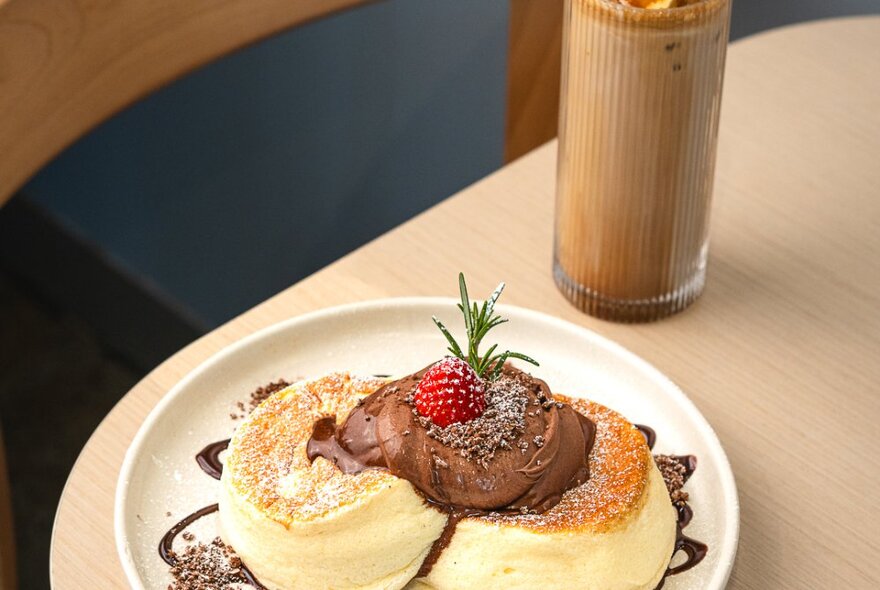 Looking across a table at souffle-style fluffy pancakes on a plate with chocolate sauce and a strawberry, a chocolate drink in the background. 