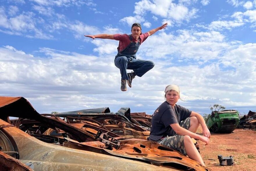 Two hound men in caps look in the desert next to a graveyard of rusted cars, one sitting and the other leaping in the air. 