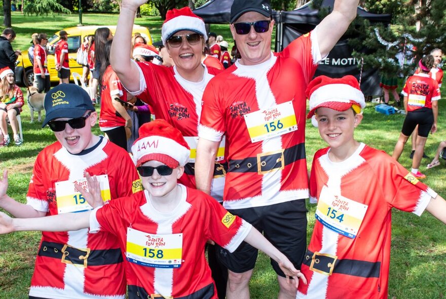 A group of two adults and three children wearing Santa red t-shirts, smiling and with their arms raised in the air, in a park, all wearing numbered running bibs.