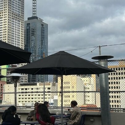 People seated on a rooftop bar under umbrellas behind wire fence, city buildings and grey sky.