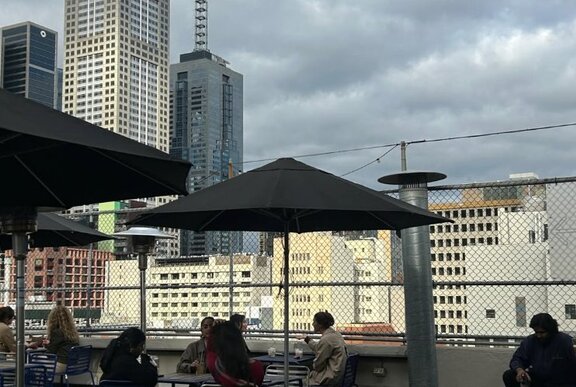 People seated on a rooftop bar under umbrellas behind wire fence, city buildings and grey sky.