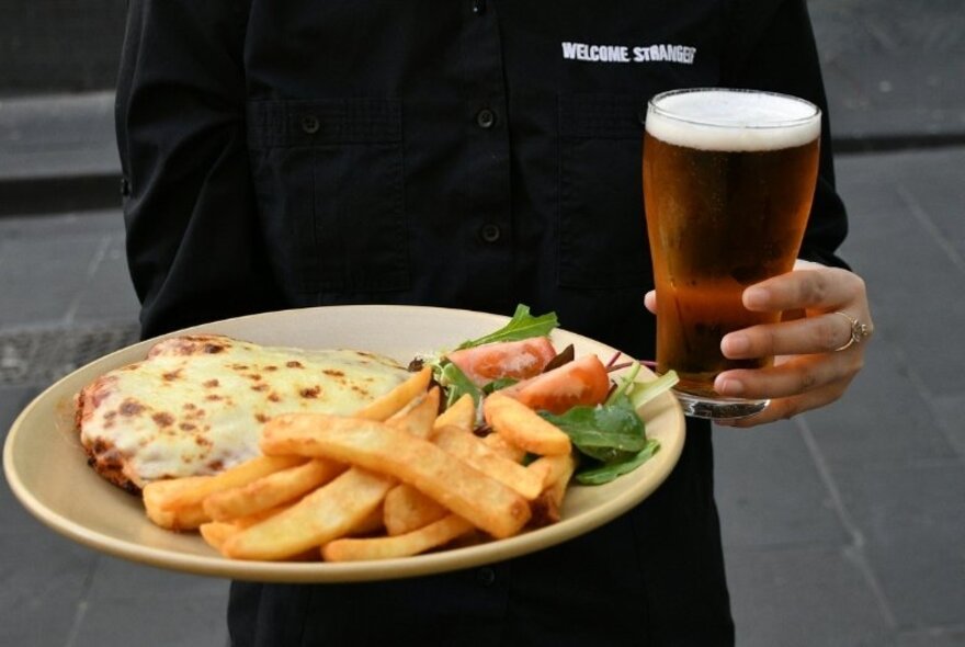 The torso of a waiter dressed in black holding a plate of chicken parmigiana and a glass of beer. 