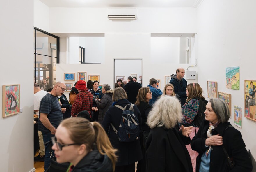 A crowd of people in a gallery space, looking at small, colourful artworks displayed on the white walls.