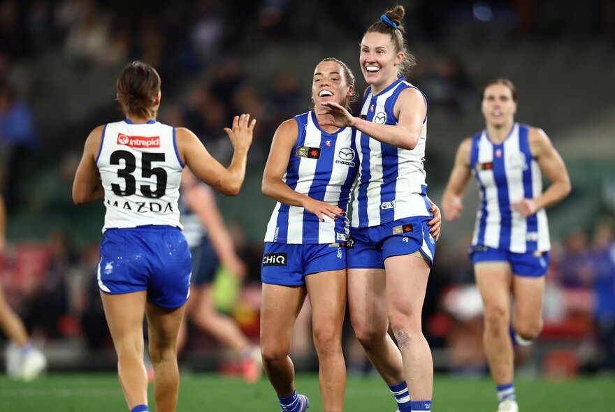 North Melbourne AFLW players embrace in celebration on the football field.