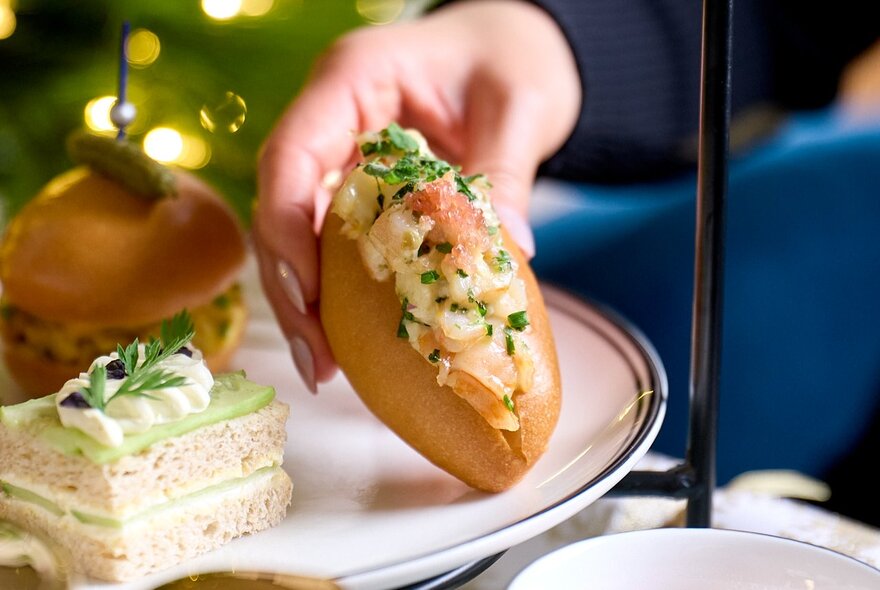 A hand selecting a small lobster roll from a plate as part of a high tea.