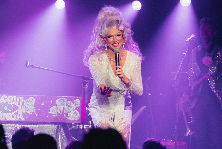 A smiling drag queen with big hair performing on stage with purple lighting. 