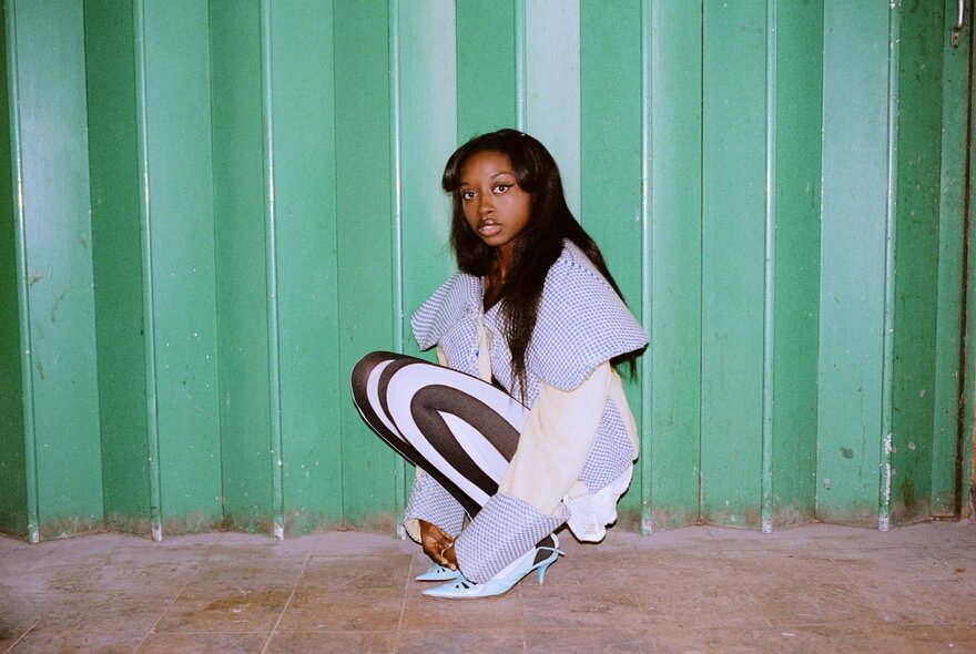 Jazz musician, Amy Gadiaga, crouching in front of a green metal background, wearing a black and white outfit.