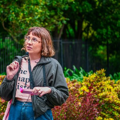 A woman with a headset and portable microphone, standing in front of a lush garden.