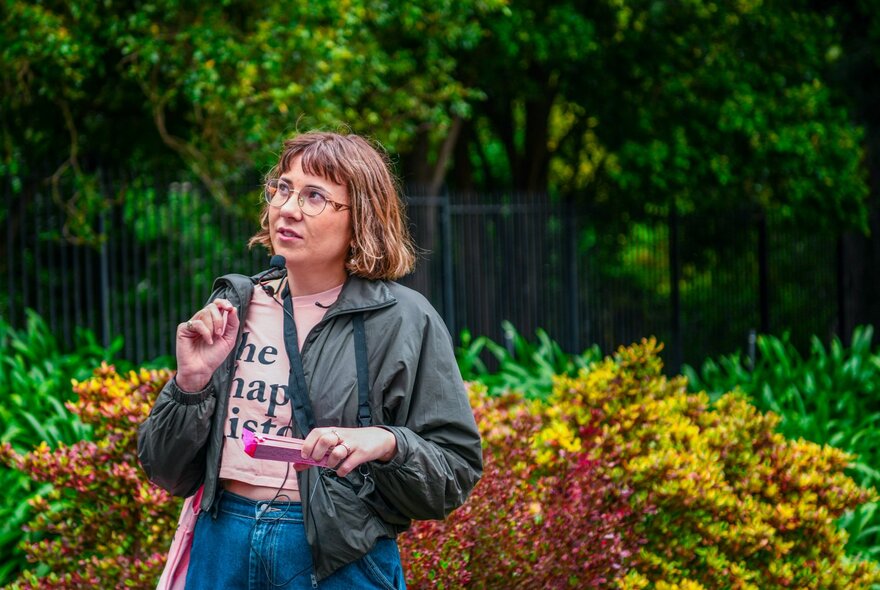 A woman with a headset and portable microphone, standing in front of a lush garden.