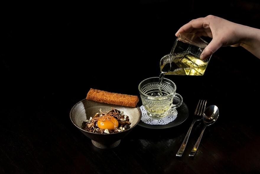 Person pouring a clear, yellow liquid into a glass cup and a small bowl of food with a raw egg yolk.