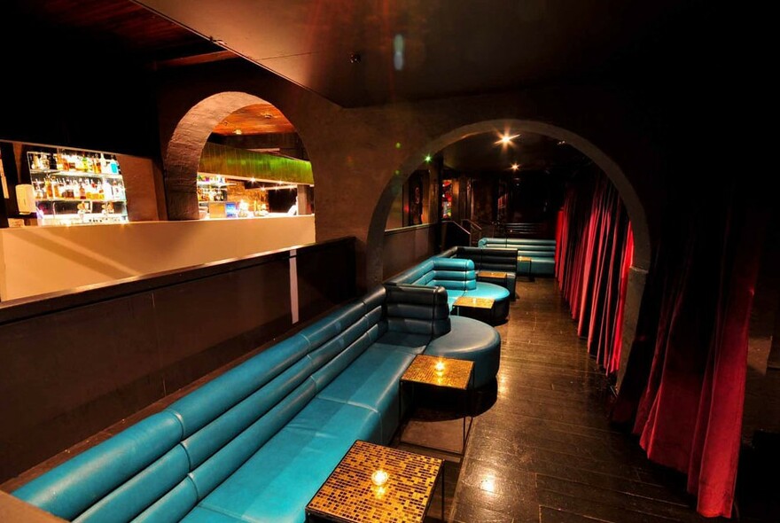 Turquoise banquette seating and tables in long room with arched doorway, red curtains and backlit bar.