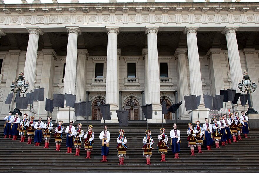 A large group of people in Ukrainian national costume lined up in a V row formation, on the steps of the Parliament of Victoria, with the facade and columns of the building in the background. 