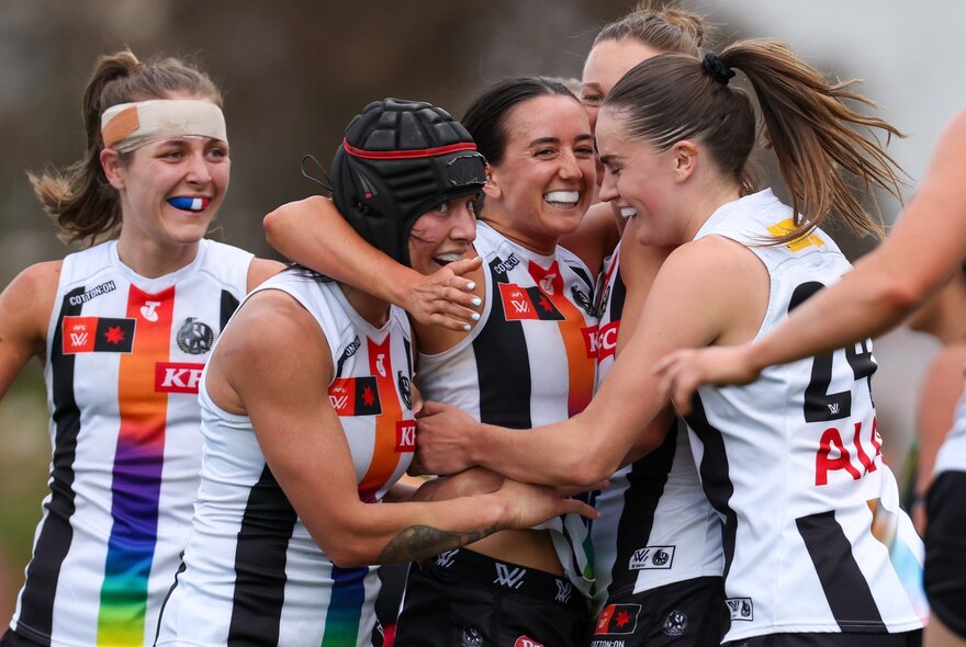 Four Collingwood Magpies AFLW players celebrating together in a group hug during a game.