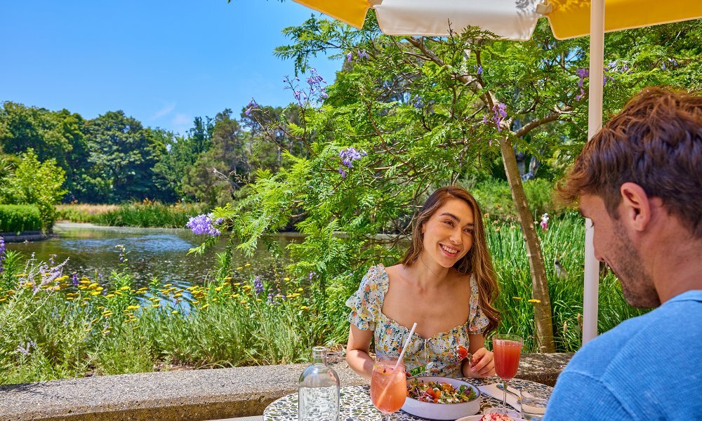 A couple dining on colourful brunch dishes and cocktails beside a lake in a sunny garden.