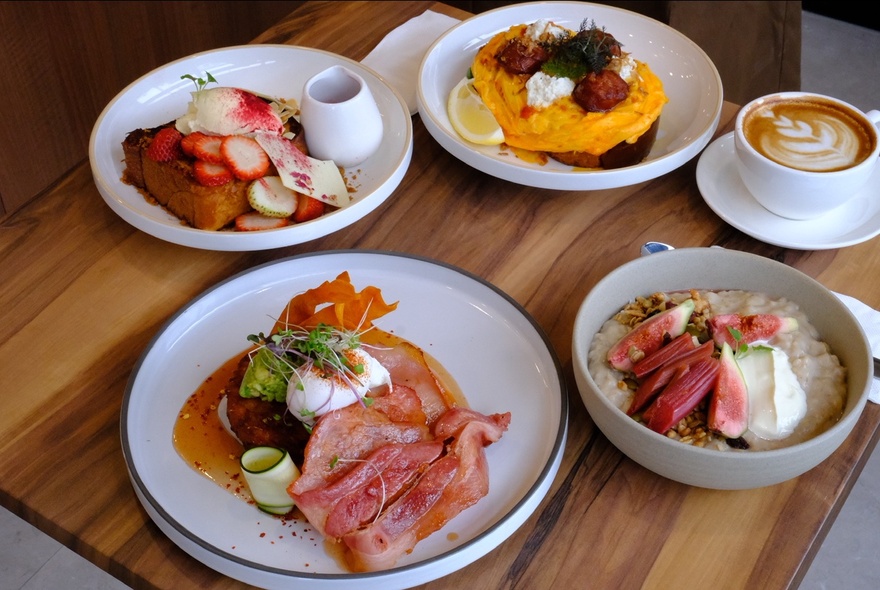 Assorted dishes on a table top including a bacon and poached egg dish, berry French toast, loaded scrambled eggs, a  bowl of porridge and fruits, paired with a café latte.