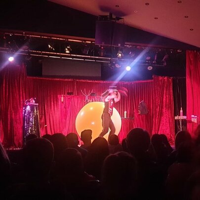Two people performing on a small stage in front of a red curtain with a seated audience watching. 