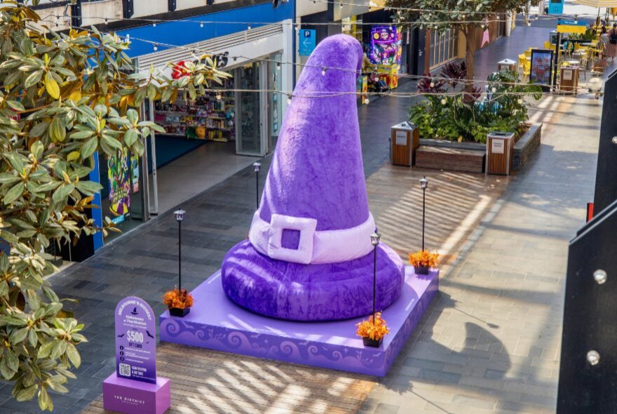 A large, plush purple witch or wizard hat set up as a Halloween display on a platform in a semi-outdoor shopping walkway at District Docklands.