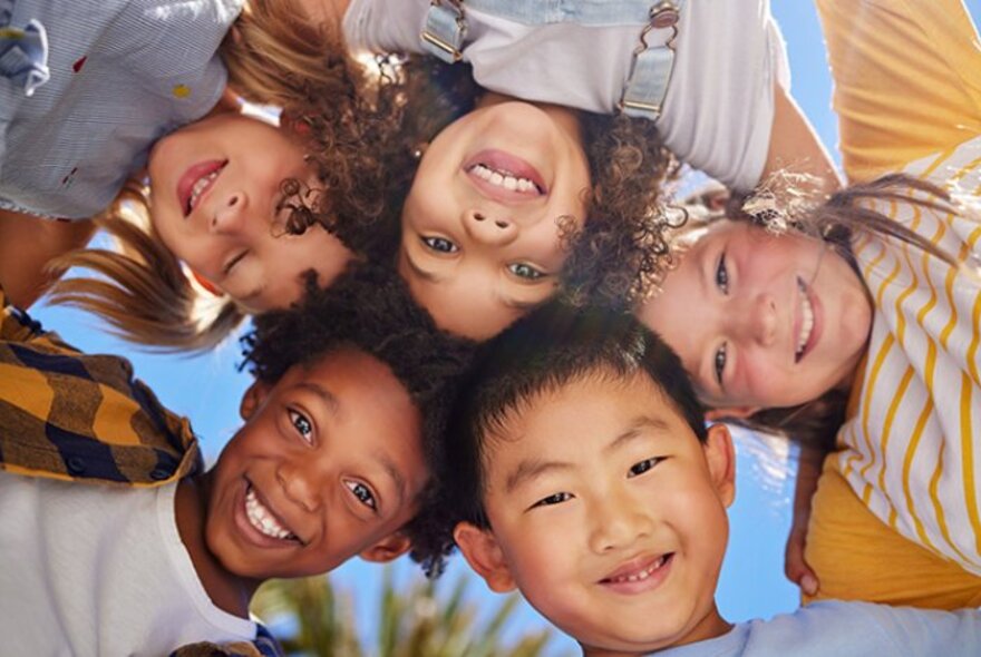 Five young children with their heads together smiling down at the camera, blue sky behind them.