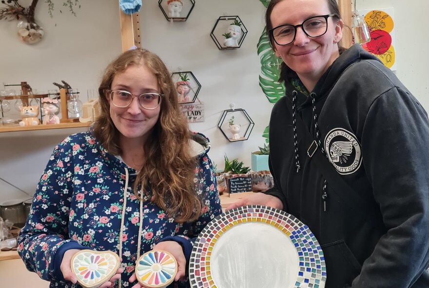 Workshop class participants smiling in a studio setting, holding up mosaic coasters and platters they have made.