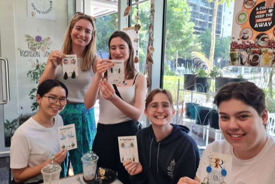 Five smiling workshop participants seated at a table with craft materials, proudly holding up jewellery they have made.
