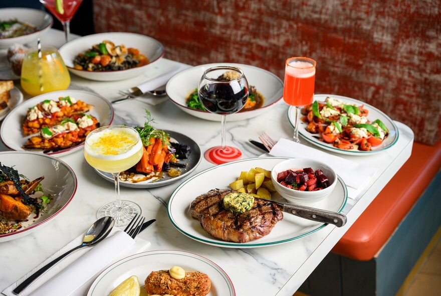 A spread of delicious food on a marble table, alongside cocktails and cutlery.