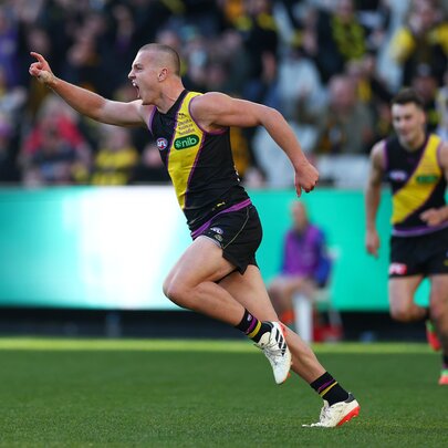 A Richmond AFL player celebrating on the grassy field, with his arm raised and finger pointing skywards, blurred fans in the stadium behind him.
