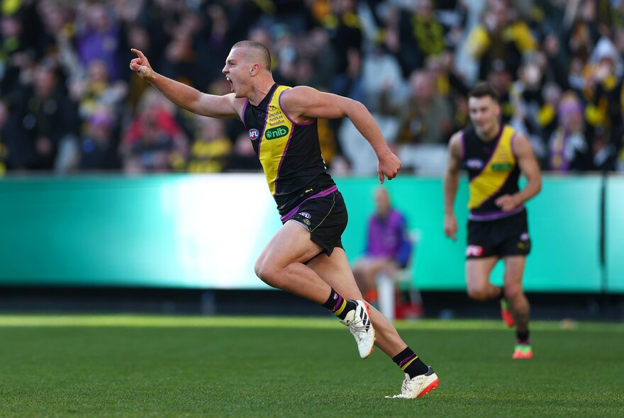 A Richmond AFL player celebrating on the grassy field, with his arm raised and finger pointing skywards, blurred fans in the stadium behind him.