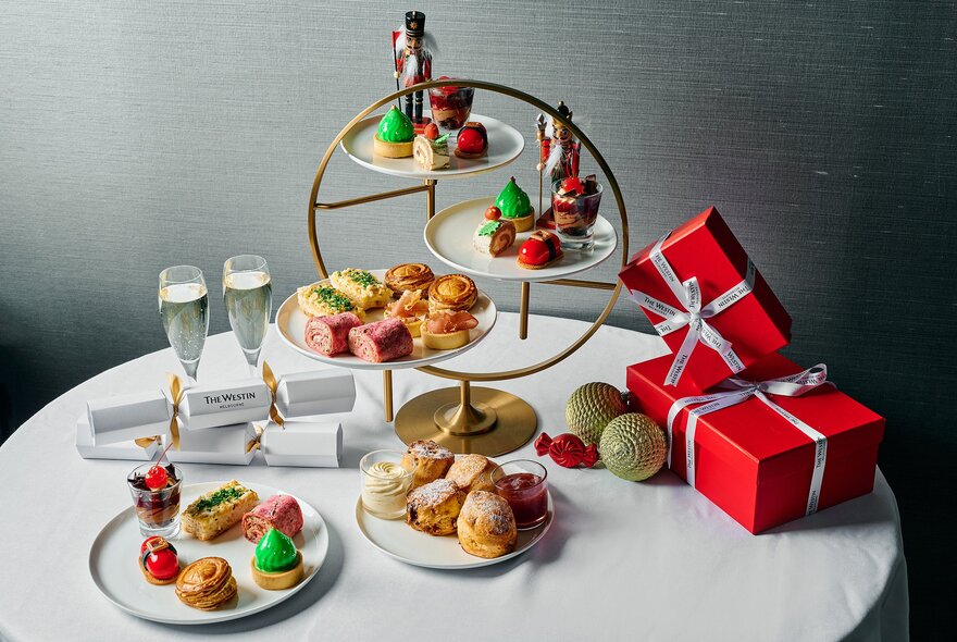 An array of Christmas-themed High Tea sweet and savoury treats on a white table alongside champagne glasses and two red-wrapped presents.