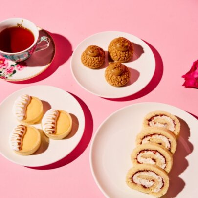 A pink table with three plates of biscuits and treats and a cup of black tea on a saucer, a rose on the table. 