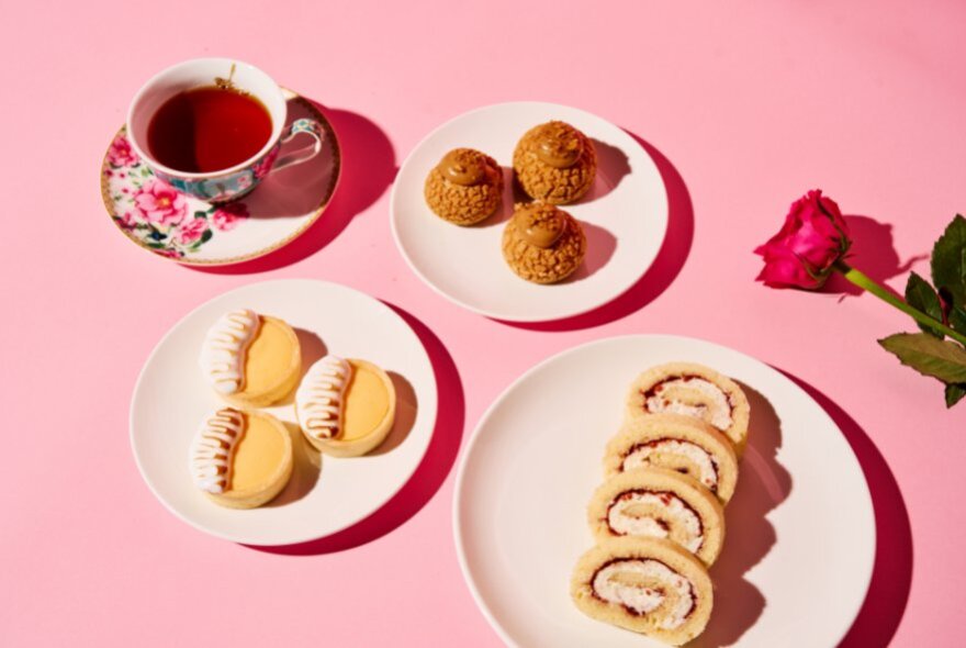 A pink table with three plates of biscuits and treats and a cup of black tea on a saucer, a rose on the table. 