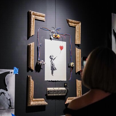 A visitor viewing Banksy’s Girl with Balloon artwork on a wall in a gallery space, rigged with a partial frame shredder mechanism, displayed as part of the Banksy Limitless exhibition.