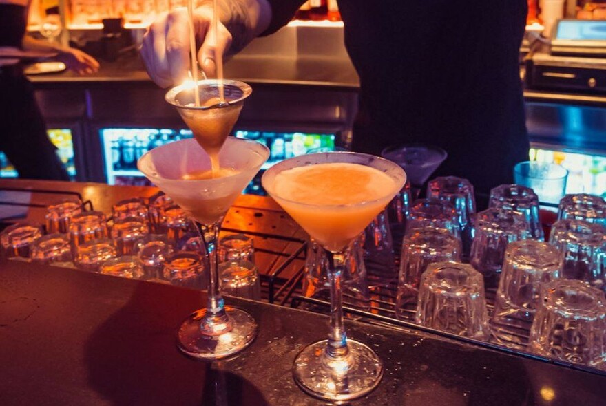 Bartender pouring two drinks into cocktail glasses on a bar counter.